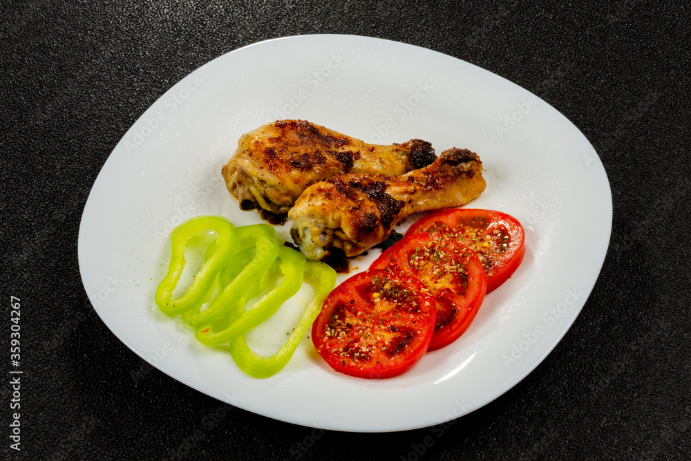 Tasty and crispy fried chicken legs, tomatoes, sweet pepper in a white plate on a black background.