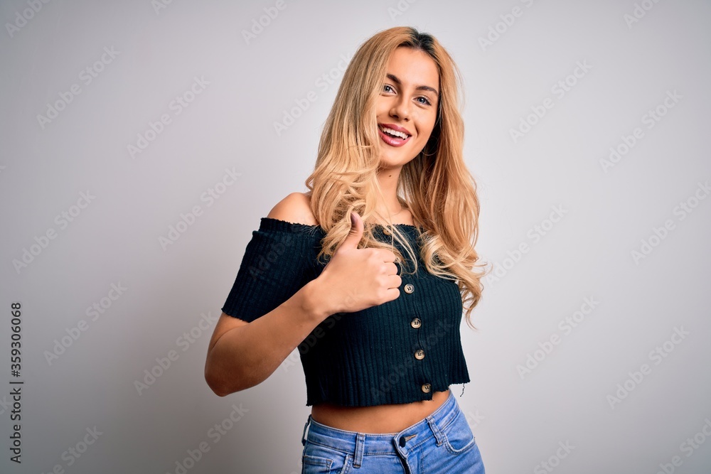 Young beautiful blonde woman wearing casual t-shirt standing over isolated white background doing happy thumbs up gesture with hand. Approving expression looking at the camera showing success. Young beautiful blonde woman wearing casual t-shirt standing over isolated white background doing happy thumbs up gesture with hand. Approving expression looking at the camera showing success.