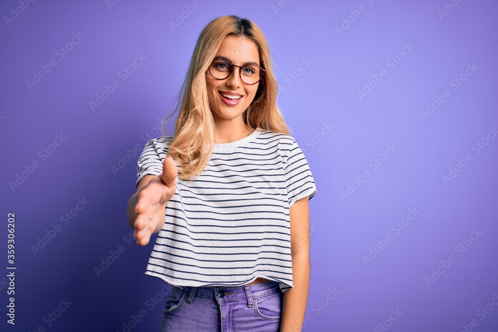 Young beautiful blonde woman wearing striped t-shirt and glasses over purple background smiling friendly offering handshake as greeting and welcoming. Successful business. Young beautiful blonde woman wearing striped t-shirt and glasses over purple background smiling friendly offering handshake as greeting and welcoming. Successful business.