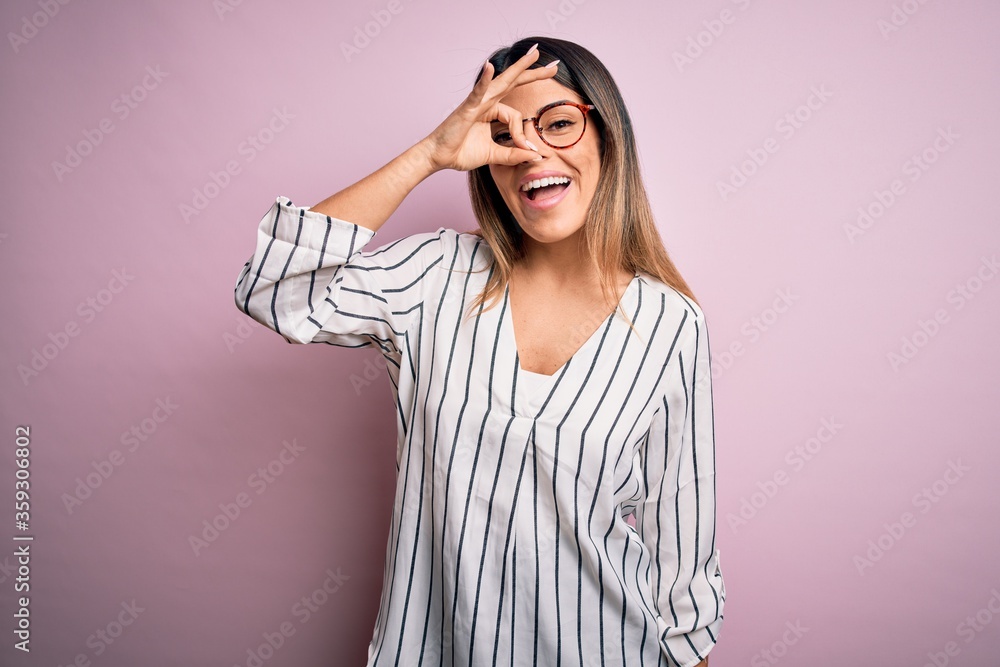 Young beautiful woman wearing casual striped t-shirt and glasses over pink background doing ok gesture with hand smiling, eye looking through fingers with happy face. Young beautiful woman wearing casual striped t-shirt and glasses over pink background doing ok gesture with hand smiling, eye looking through fingers with happy face.