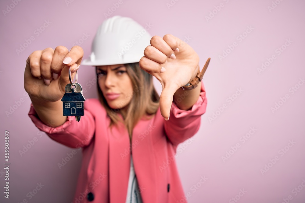 Young beautiful brunette architect woman wearing safety helmet hoding key of house with angry face, negative sign showing dislike with thumbs down, rejection concept Young beautiful brunette architect woman wearing safety helmet hoding key of house with angry face, negative sign showing dislike with thumbs down, rejection concept