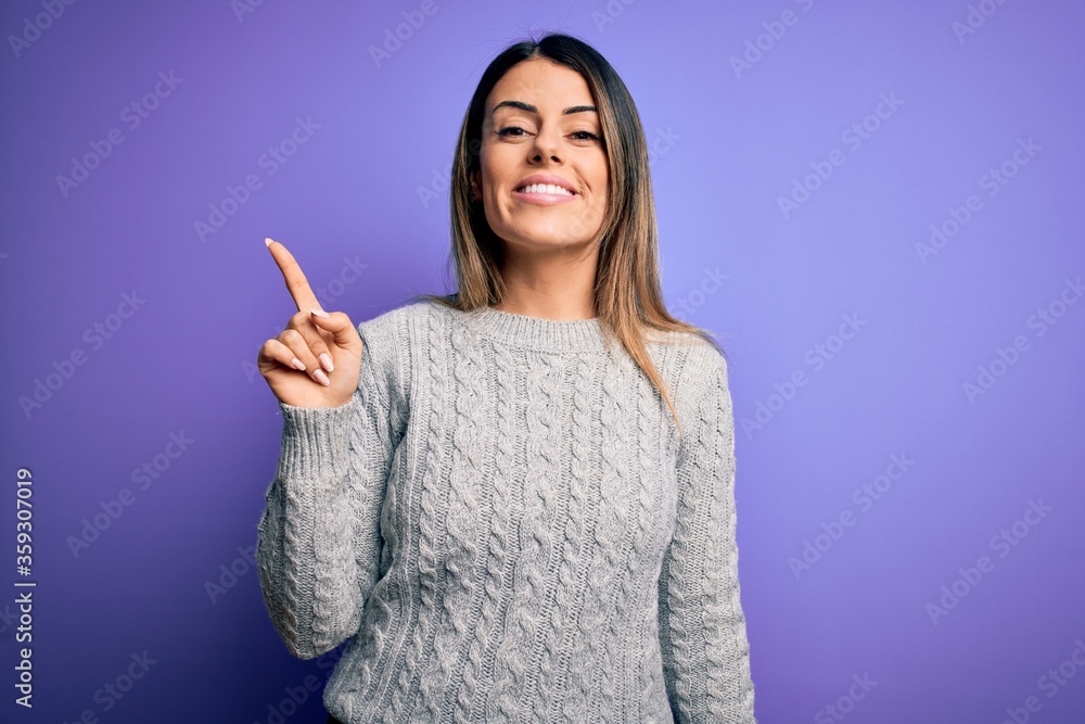 Young beautiful woman wearing casual sweater standing over isolated purple background showing and pointing up with finger number one while smiling confident and happy. Young beautiful woman wearing casual sweater standing over isolated purple background showing and pointing up with finger number one while smiling confident and happy.