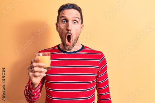 Young handsome man drinking glass of healthy orange juice over isolated yellow background scared and amazed with open mouth for surprise, disbelief face