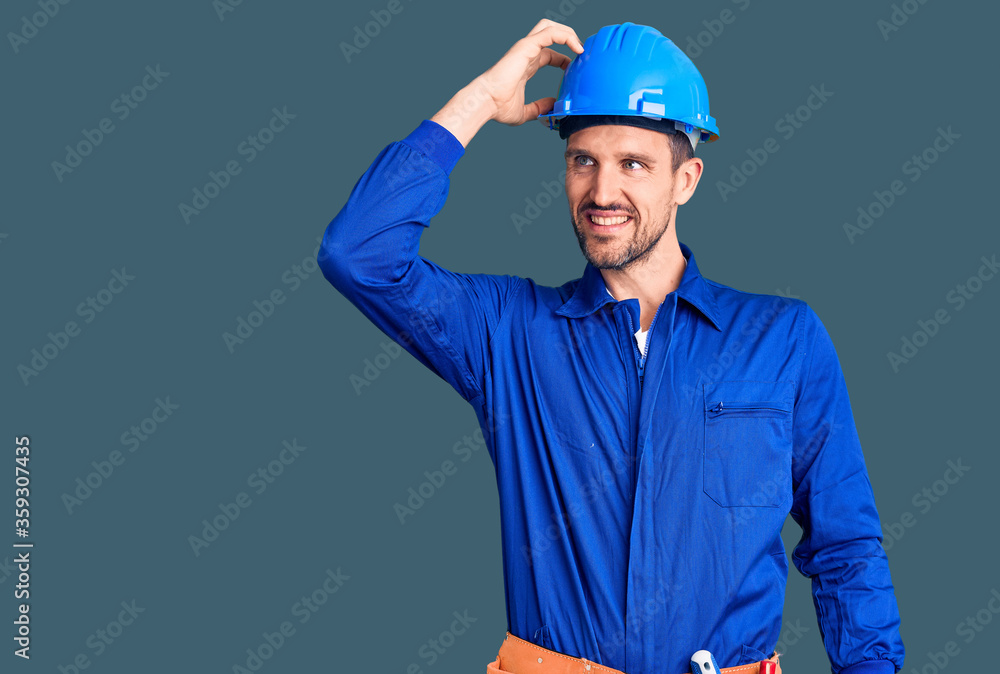 Young handsome man wearing worker uniform and hardhat smiling confident touching hair with hand up gesture, posing attractive and fashionable