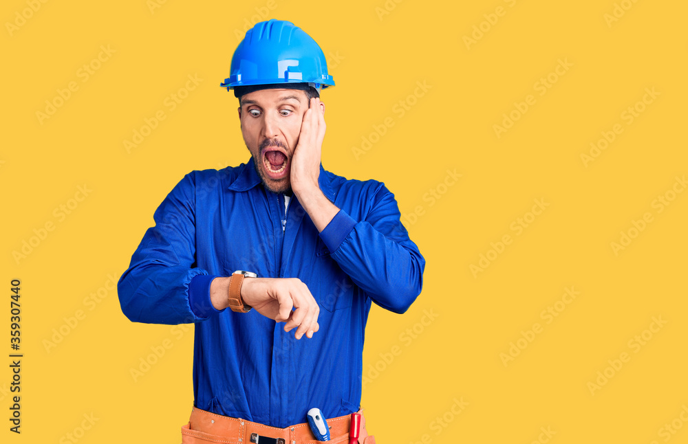 Young handsome man wearing worker uniform and hardhat amazed and surprised looking up and pointing with fingers and raised arms.