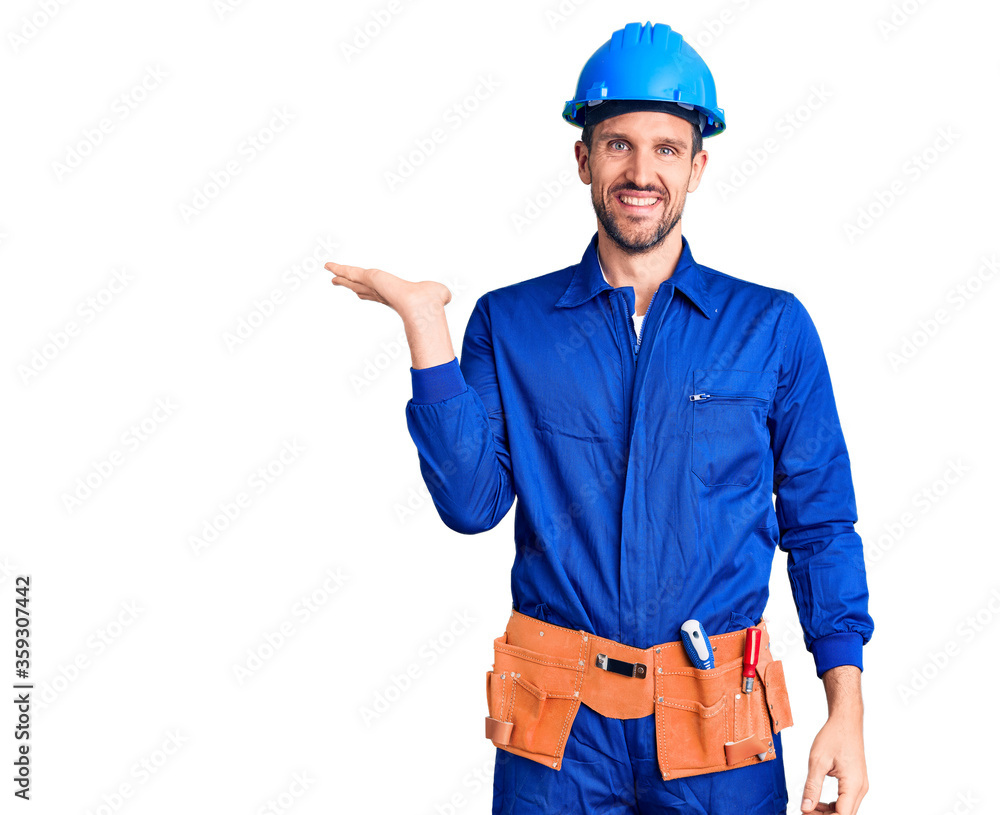 Young handsome man wearing worker uniform and hardhat smiling cheerful presenting and pointing with palm of hand looking at the camera.