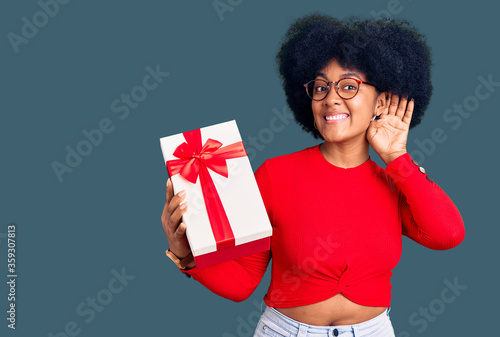 Young african american girl holding gift smiling with hand over ear listening an hearing to rumor or gossip. deafness concept.