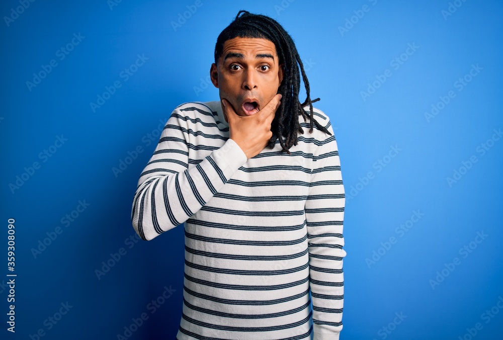 Young handsome african american afro man with dreadlocks wearing casual striped sweater Looking fascinated with disbelief, surprise and amazed expression with hands on chin Young handsome african american afro man with dreadlocks wearing casual striped sweater Looking fascinated with disbelief, surprise and amazed expression with hands on chin