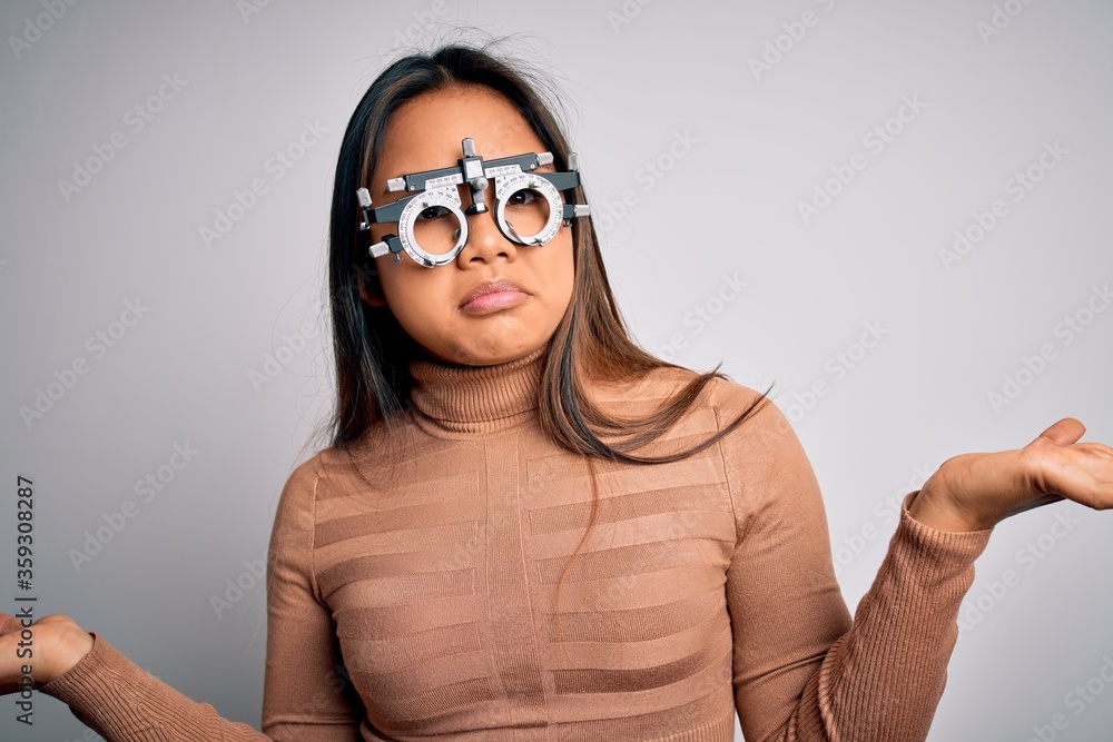 Young asian optical girl controlling eyesight using optometry glasses over white background clueless and confused expression with arms and hands raised. Doubt concept. Young asian optical girl controlling eyesight using optometry glasses over white background clueless and confused expression with arms and hands raised. Doubt concept.