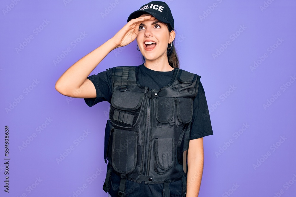 Young police woman wearing security bulletproof vest uniform over purple background very happy and smiling looking far away with hand over head. Searching concept. Young police woman wearing security bulletproof vest uniform over purple background very happy and smiling looking far away with hand over head. Searching concept.