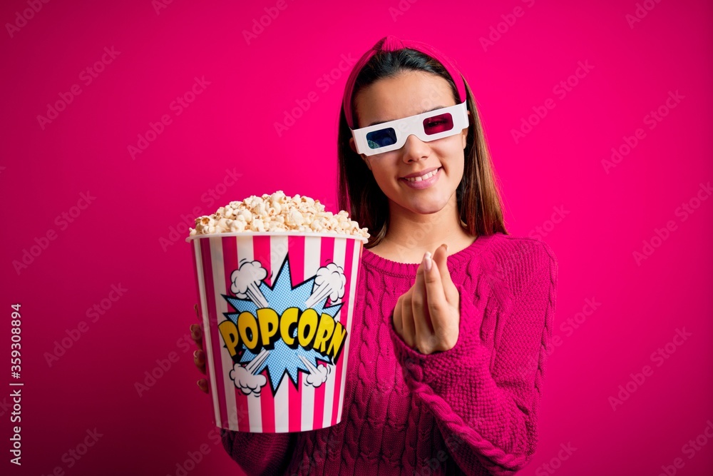 Young beautiful girl watching movie using 3d glasses eating box with popcorns doing money gesture with hands, asking for salary payment, millionaire business