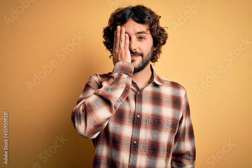 Young handsome man with beard wearing casual shirt standing over yellow background covering one eye with hand, confident smile on face and surprise emotion.