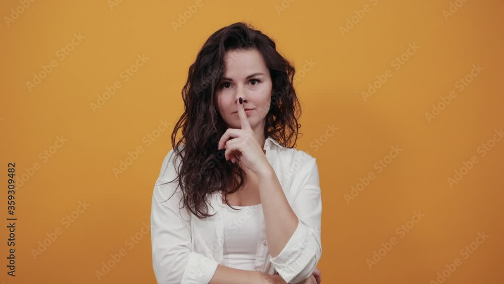 Index finger on lips, silence gesture, shhh quiet, asks for voicelessness forefinger at the mouth. Young attractive woman, dressed white blouse, with brown eyes, curly hair, yellow background Index finger on lips, silence gesture, shhh quiet, asks for voicelessness forefinger at the mouth. Young attractive woman, dressed white blouse, with brown eyes, curly hair, yellow background
