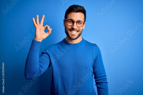 Young handsome man with beard wearing casual sweater and glasses over blue background smiling positive doing ok sign with hand and fingers. Successful expression.
