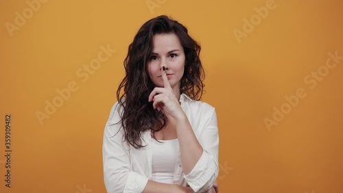 Index finger on lips, silence gesture, shhh quiet, asks for voicelessness forefinger at the mouth. Young attractive woman, dressed white blouse, with brown eyes, curly hair, yellow background photo