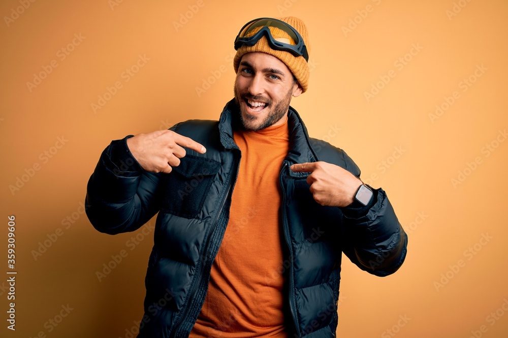 Young handsome skier man with beard wearing snow sportswear and ski goggles looking confident with smile on face, pointing oneself with fingers proud and happy.