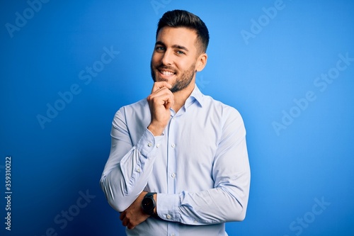 Young handsome man wearing elegant shirt standing over isolated blue background looking confident at the camera with smile with crossed arms and hand raised on chin. Thinking positive.