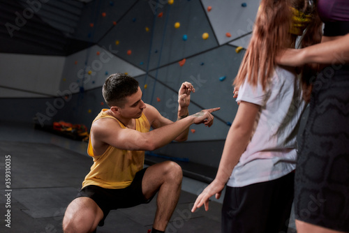 Male instructor helping active little girl going to climb, standing against artificial training climbing wall