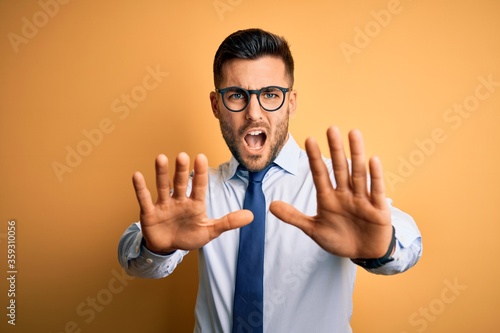 Young handsome businessman wearing tie and glasses standing over yellow background doing stop gesture with hands palms, angry and frustration expression © Krakenimages.com