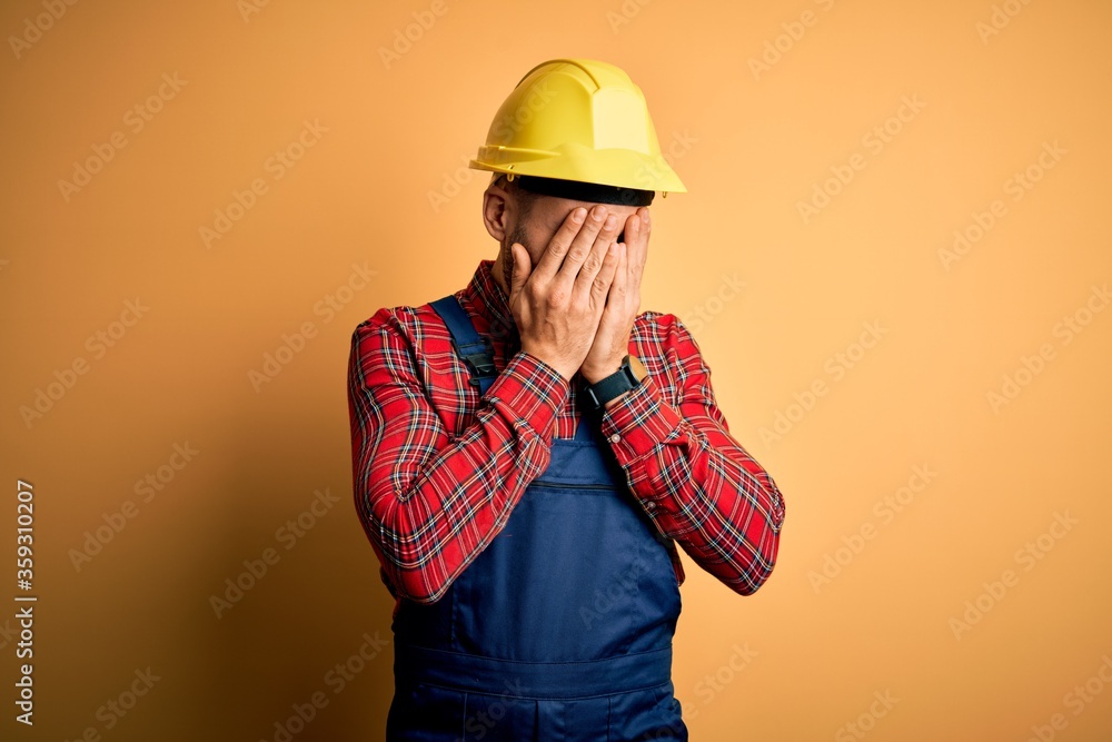 Young builder man wearing construction uniform and safety helmet over yellow isolated background with sad expression covering face with hands while crying. Depression concept. Young builder man wearing construction uniform and safety helmet over yellow isolated background with sad expression covering face with hands while crying. Depression concept.