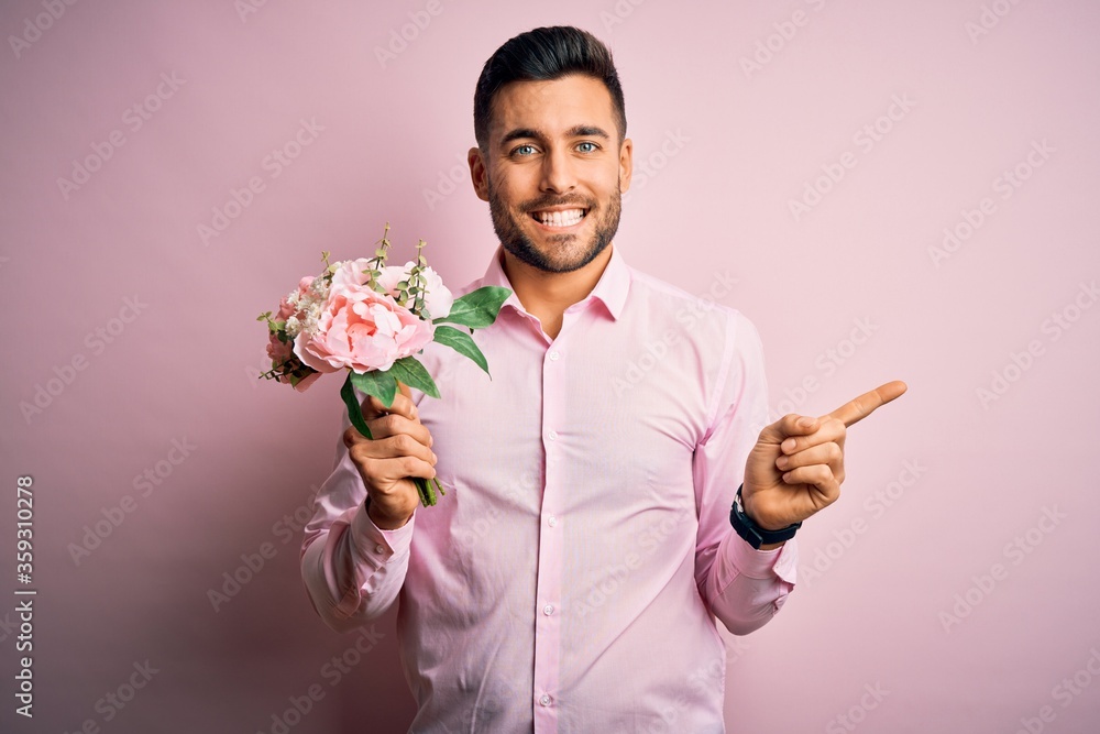 Young romatic man holding bouquet of spring flowers over pink isolatd background very happy pointing with hand and finger to the side Young romatic man holding bouquet of spring flowers over pink isolatd background very happy pointing with hand and finger to the side