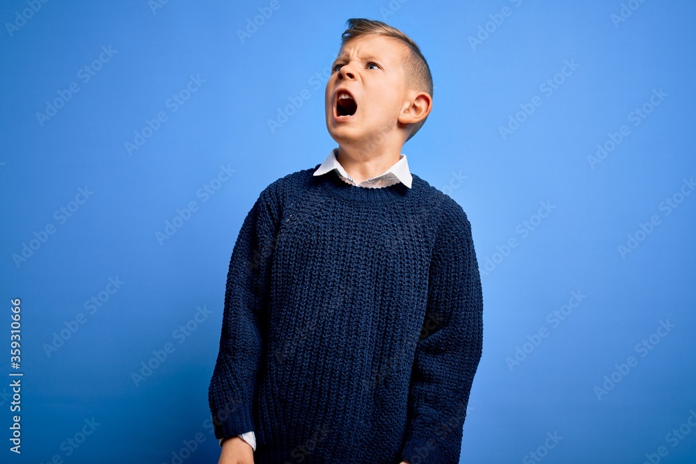 Young little caucasian kid with blue eyes wearing winter sweater over blue background crazy and mad shouting and yelling with aggressive expression and arms raised. Frustration concept. Young little caucasian kid with blue eyes wearing winter sweater over blue background crazy and mad shouting and yelling with aggressive expression and arms raised. Frustration concept.