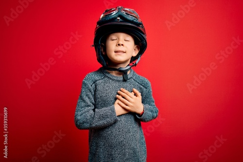Young little caucasian kid wearing vintage biker motorcycle helmet and googles over red background smiling with hands on chest with closed eyes and grateful gesture on face. Health concept.