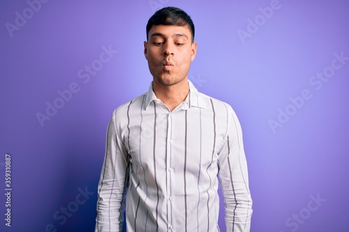 Young handsome hispanic man wearing elegant business shirt standing over purple background making fish face with lips, crazy and comical gesture. Funny expression.