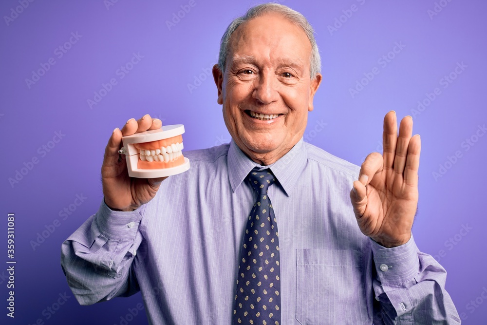 Grey haired senior man holding orthodontic prosthesis denture over purple background doing ok sign with fingers, excellent symbol Grey haired senior man holding orthodontic prosthesis denture over purple background doing ok sign with fingers, excellent symbol