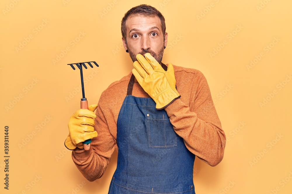 Young handsome man wearing gardener apron and gloves holding rake covering mouth with hand, shocked and afraid for mistake. surprised expression Young handsome man wearing gardener apron and gloves holding rake covering mouth with hand, shocked and afraid for mistake. surprised expression