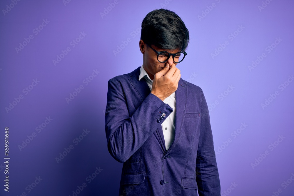 Young handsome business man wearing jacket and glasses over isolated purple background smelling something stinky and disgusting, intolerable smell, holding breath with fingers on nose. Bad smell Young handsome business man wearing jacket and glasses over isolated purple background smelling something stinky and disgusting, intolerable smell, holding breath with fingers on nose. Bad smell