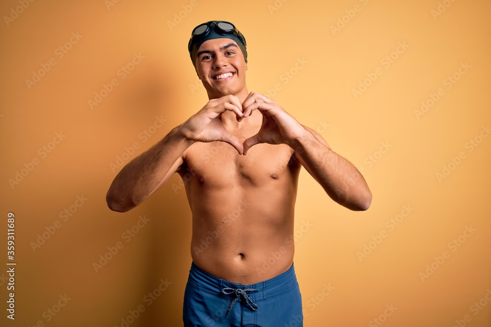 Young handsome man shirtless wearing swimsuit and swim cap over isolated yellow background smiling in love doing heart symbol shape with hands. Romantic concept.
