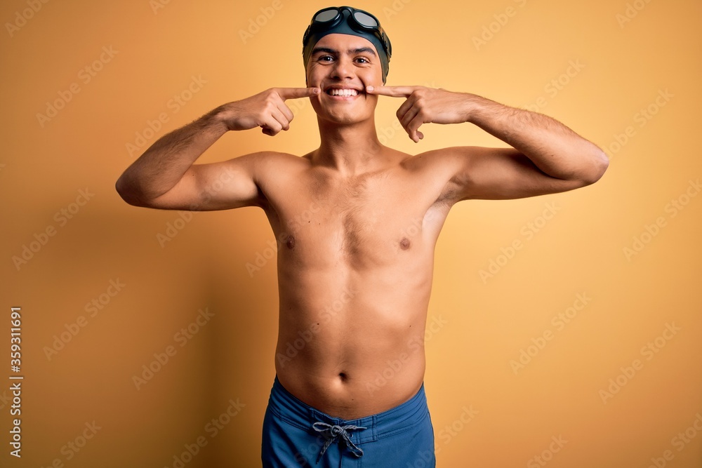 Young handsome man shirtless wearing swimsuit and swim cap over isolated yellow background smiling cheerful showing and pointing with fingers teeth and mouth. Dental health concept.