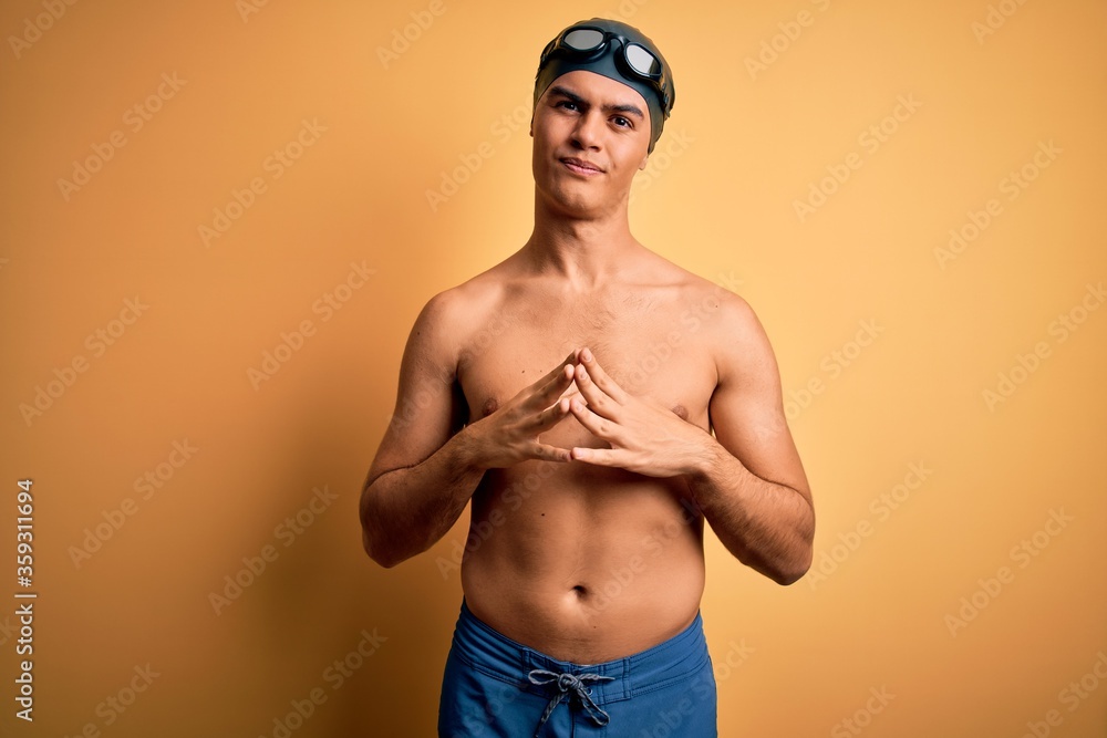 Young handsome man shirtless wearing swimsuit and swim cap over isolated yellow background Hands together and fingers crossed smiling relaxed and cheerful. Success and optimistic