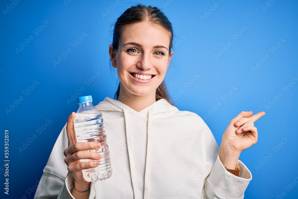 Young beautiful redhead woman doing sport drinking bottle of water over blue background very happy pointing with hand and finger to the side Young beautiful redhead woman doing sport drinking bottle of water over blue background very happy pointing with hand and finger to the side