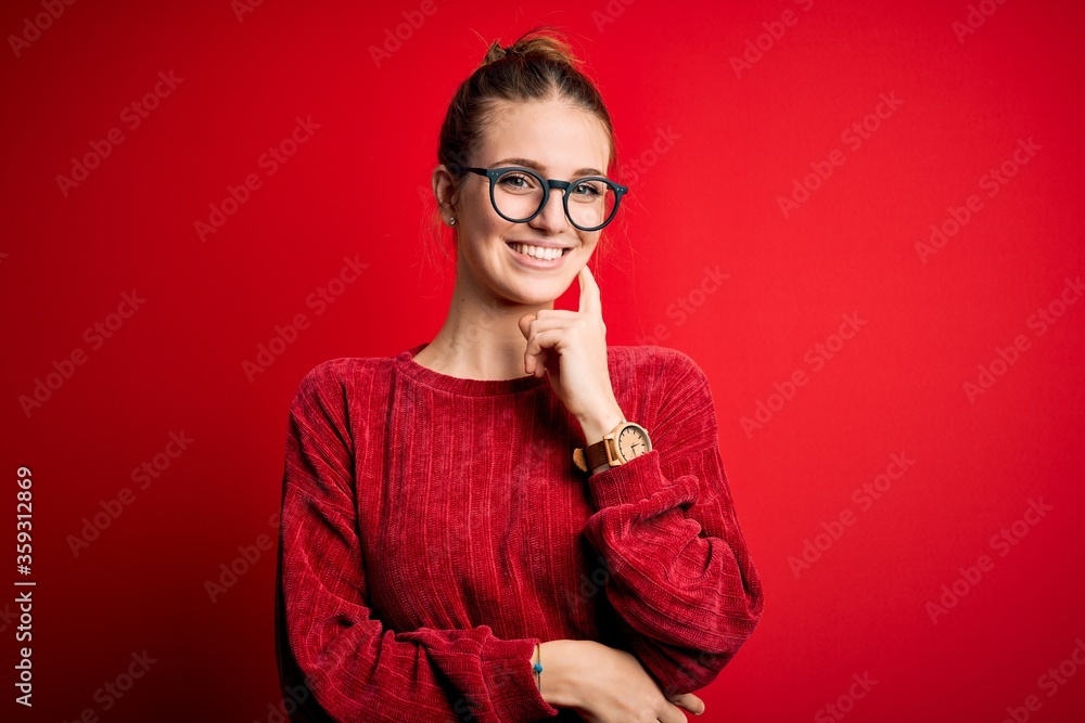 Young beautiful redhead woman wearing casual sweater over isolated red background looking confident at the camera smiling with crossed arms and hand raised on chin. Thinking positive. Young beautiful redhead woman wearing casual sweater over isolated red background looking confident at the camera smiling with crossed arms and hand raised on chin. Thinking positive.