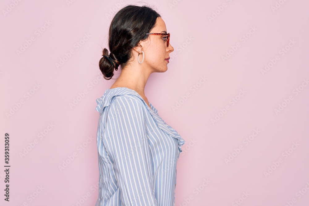 Young beautiful woman wearing casual striped shirt and glasses over pink background looking to side, relax profile pose with natural face with confident smile. Young beautiful woman wearing casual striped shirt and glasses over pink background looking to side, relax profile pose with natural face with confident smile.