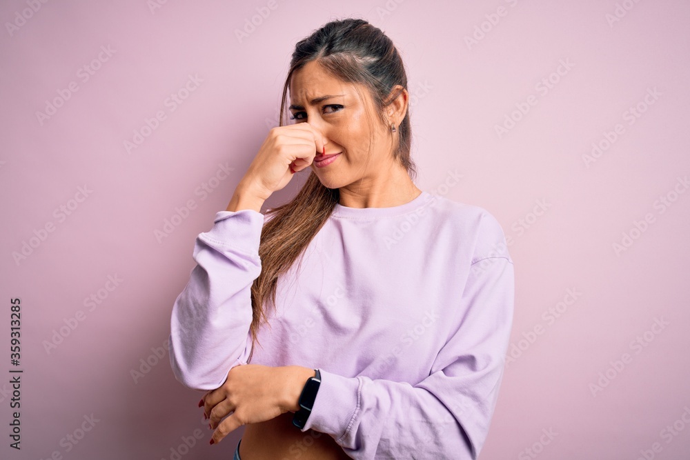 Young beautiful sport woman wearing sweatshirt over pink isolated background smelling something stinky and disgusting, intolerable smell, holding breath with fingers on nose. Bad smell Young beautiful sport woman wearing sweatshirt over pink isolated background smelling something stinky and disgusting, intolerable smell, holding breath with fingers on nose. Bad smell
