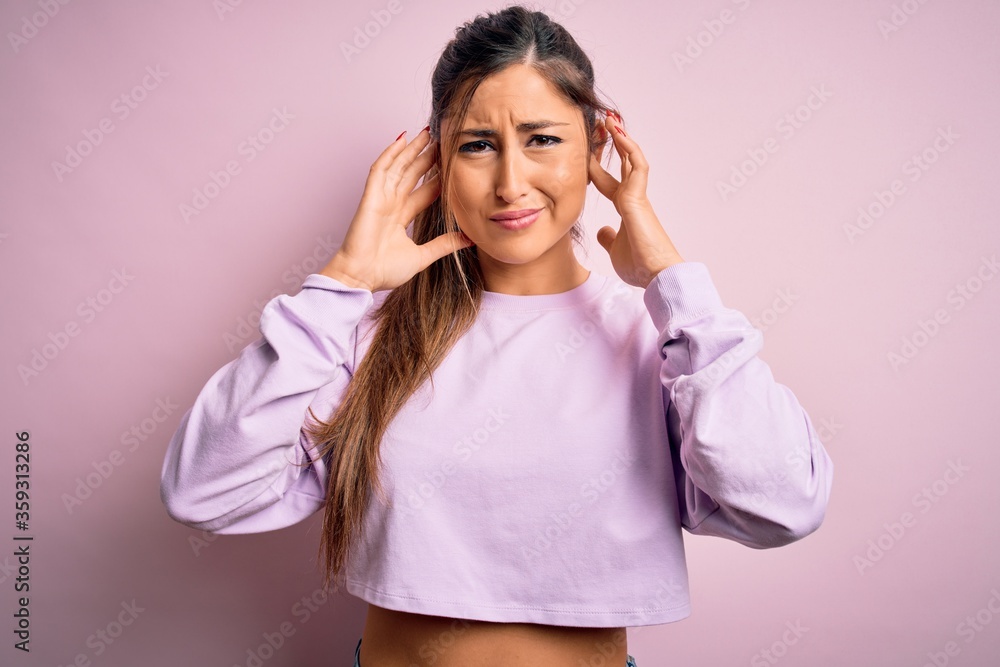 Young beautiful sport woman wearing sweatshirt over pink isolated background covering ears with fingers with annoyed expression for the noise of loud music. Deaf concept. Young beautiful sport woman wearing sweatshirt over pink isolated background covering ears with fingers with annoyed expression for the noise of loud music. Deaf concept.