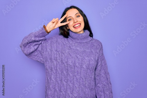 Young beautiful woman wearing casual turtleneck sweater standing over purple background Doing peace symbol with fingers over face, smiling cheerful showing victory