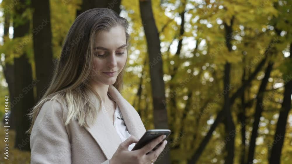 Beautiful lady in coat typing on mobile phone with cute smile at autumn park Beautiful lady in coat typing on mobile phone with cute smile at autumn park