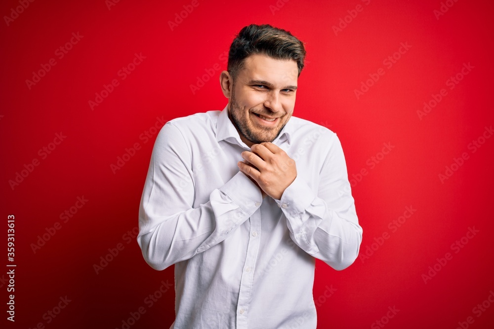 Young business man with blue eyes wearing elegant shirt standing over red isolated background laughing nervous and excited with hands on chin looking to the side Young business man with blue eyes wearing elegant shirt standing over red isolated background laughing nervous and excited with hands on chin looking to the side