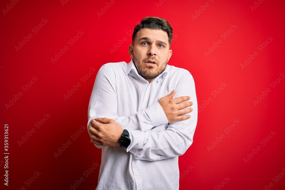 Young business man with blue eyes wearing elegant shirt standing over red isolated background shaking and freezing for winter cold with sad and shock expression on face Young business man with blue eyes wearing elegant shirt standing over red isolated background shaking and freezing for winter cold with sad and shock expression on face