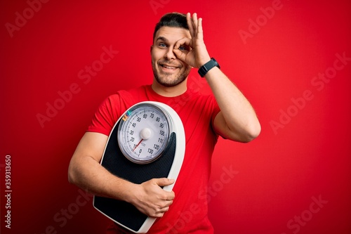 Young fitness man with blue eyes holding scale dieting for healthy weight over red background with happy face smiling doing ok sign with hand on eye looking through fingers