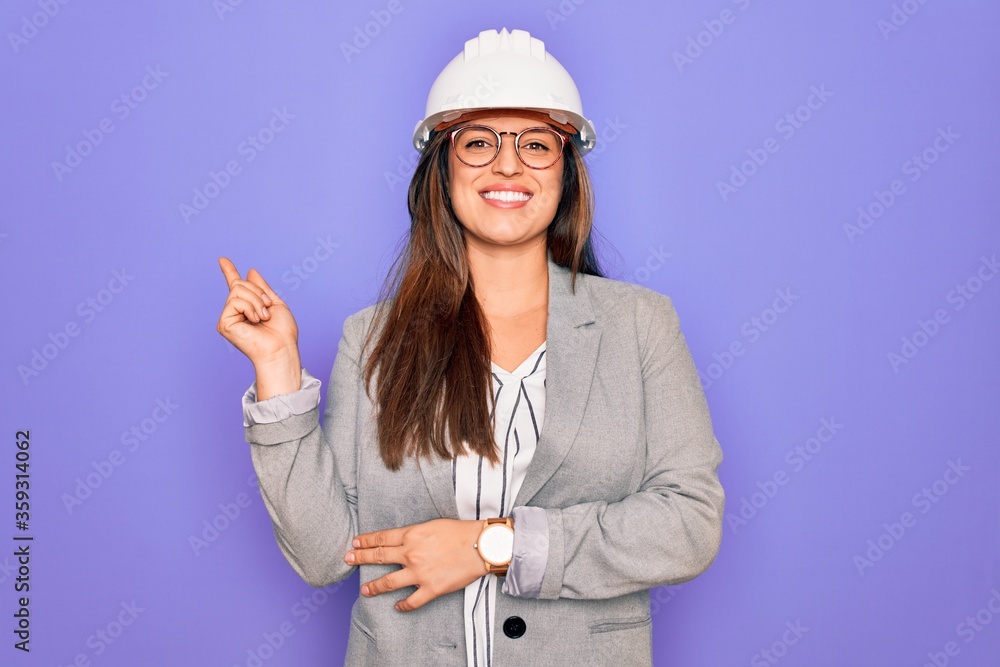 Professional woman engineer wearing industrial safety helmet over pruple background with a big smile on face, pointing with hand and finger to the side looking at the camera.