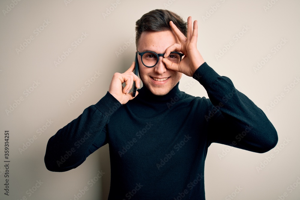 Young handsome caucasian man having a conversation talking on smartphone with happy face smiling doing ok sign with hand on eye looking through fingers Young handsome caucasian man having a conversation talking on smartphone with happy face smiling doing ok sign with hand on eye looking through fingers
