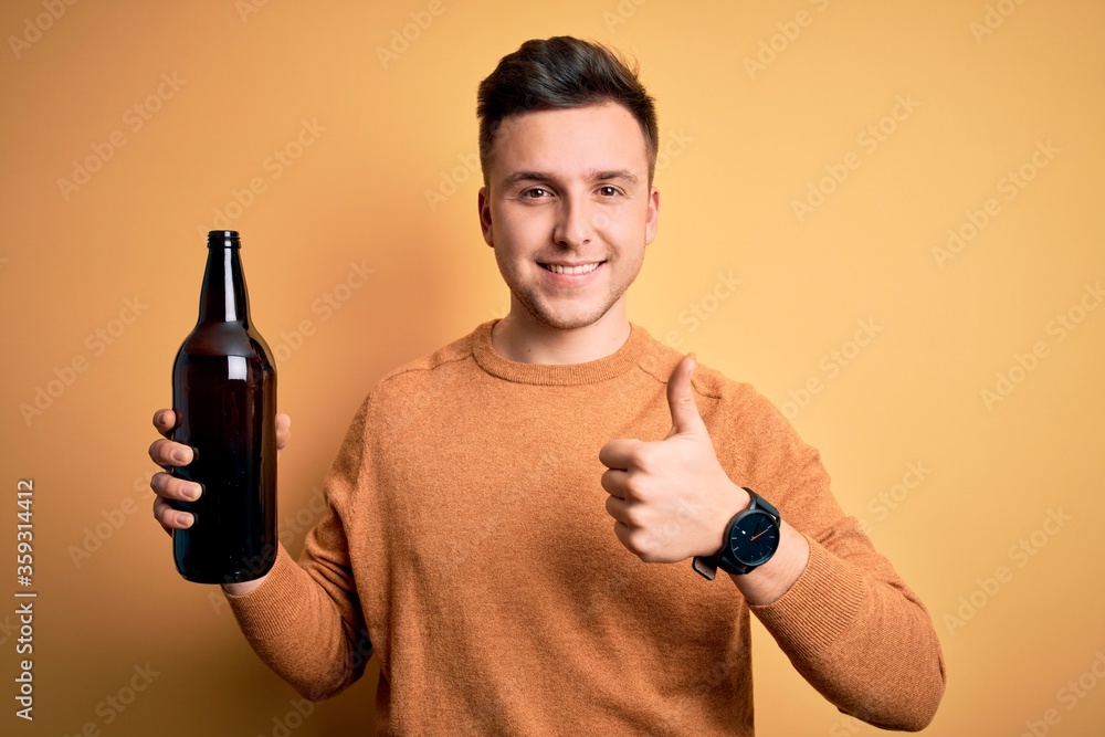 Young handsome caucasian man drinking a bottle of beer over yellow background happy with big smile doing ok sign, thumb up with fingers, excellent sign Young handsome caucasian man drinking a bottle of beer over yellow background happy with big smile doing ok sign, thumb up with fingers, excellent sign