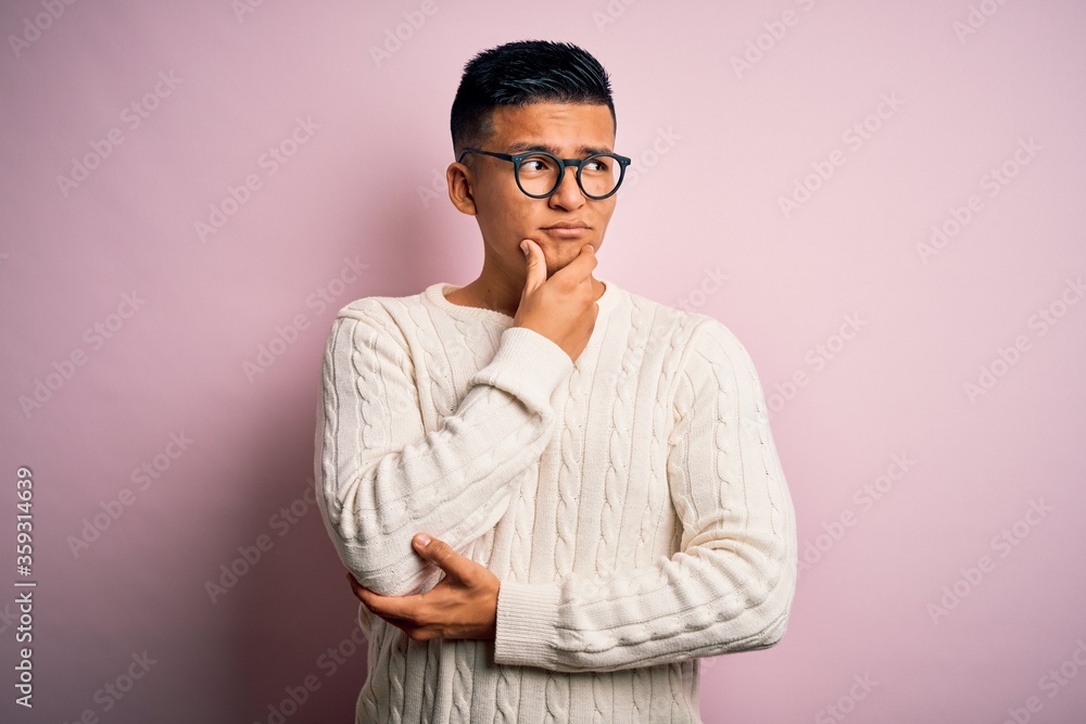 Young handsome latin man wearing white casual sweater and glasses over pink background with hand on chin thinking about question, pensive expression. Smiling with thoughtful face. Doubt concept. Young handsome latin man wearing white casual sweater and glasses over pink background with hand on chin thinking about question, pensive expression. Smiling with thoughtful face. Doubt concept.