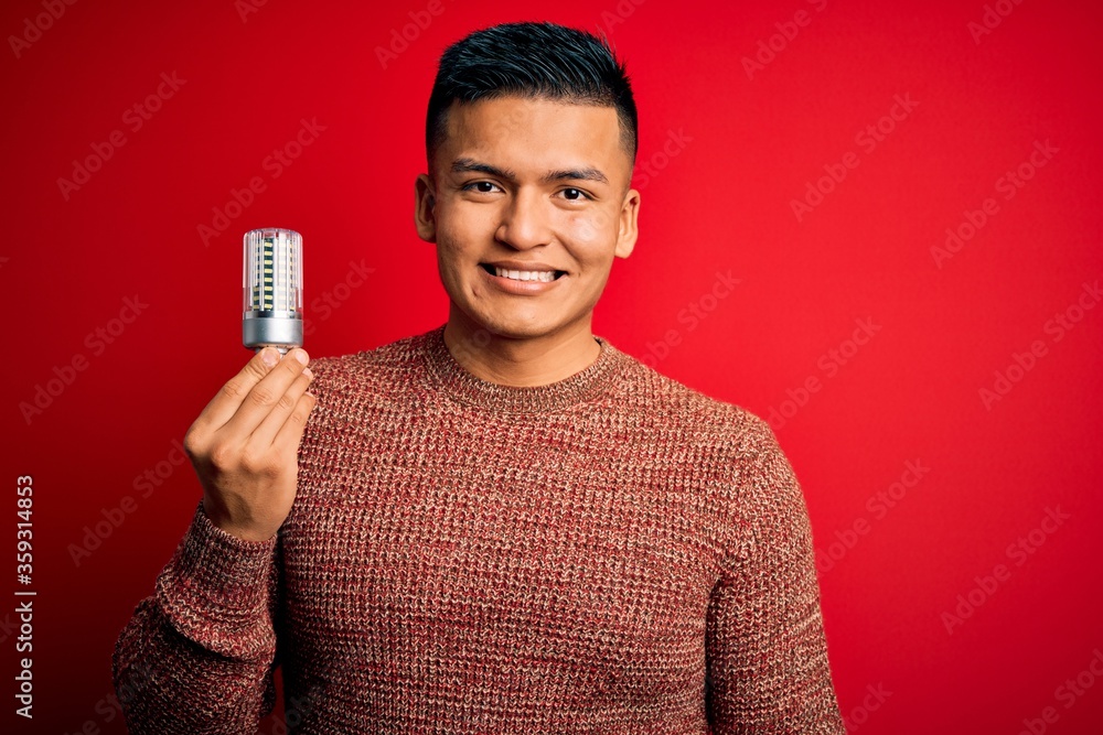 Young handsome latin man holding led lightbulb over isolated red background with a happy face standing and smiling with a confident smile showing teeth Young handsome latin man holding led lightbulb over isolated red background with a happy face standing and smiling with a confident smile showing teeth