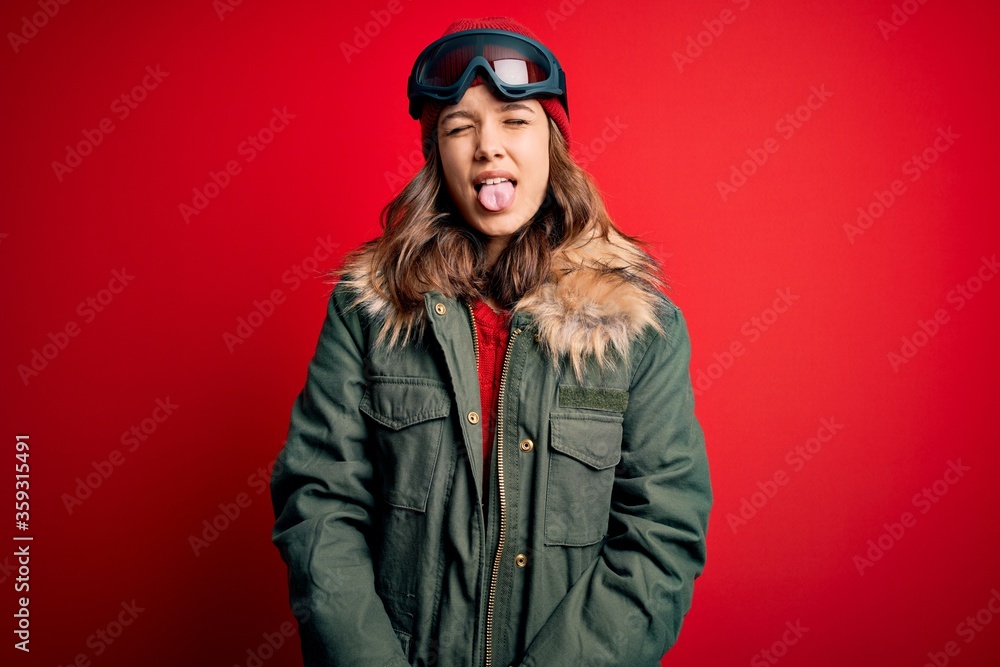 Young blonde girl wearing ski glasses and winter coat for ski weather over red background sticking tongue out happy with funny expression. Emotion concept. Young blonde girl wearing ski glasses and winter coat for ski weather over red background sticking tongue out happy with funny expression. Emotion concept.
